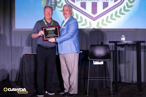 Two men on a stage exchange a plaque during an awards ceremony, with a large crest backdrop behind them; one in a gray polo, the other in a light blue blazer.