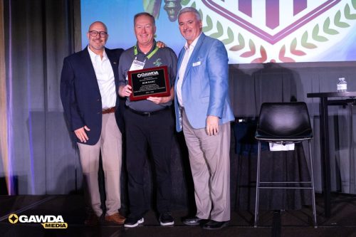 Three men on stage posing with an award plaque, center recipient smiling; GAWDA Media logo visible bottom left.
