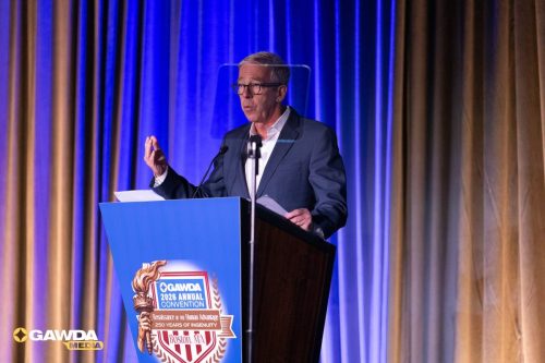 Male speaker in a gray suit and glasses delivering a speech at a podium on a stage with blue curtains during the GAWDA 2026 Annual Convention in Boston, MA (GAWDA logo visible).