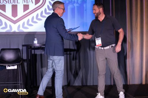 Two men shake hands on a stage during an award ceremony; one in a suit, the other wearing a polo with a badge.