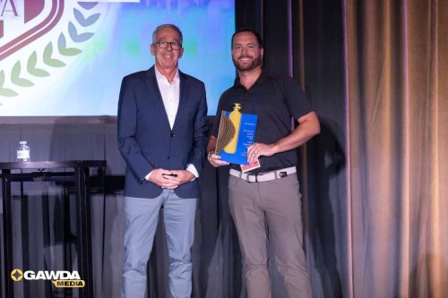 Two men stand on a stage at an awards ceremony; one in a navy blazer, the other in a dark polo holding a blue award plaque and yellow device.