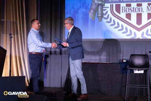 Two men in business attire shake hands on a stage during an award presentation; the man on the right hands a folder to the man on the left as a banner with a Boston crest is projected behind them.