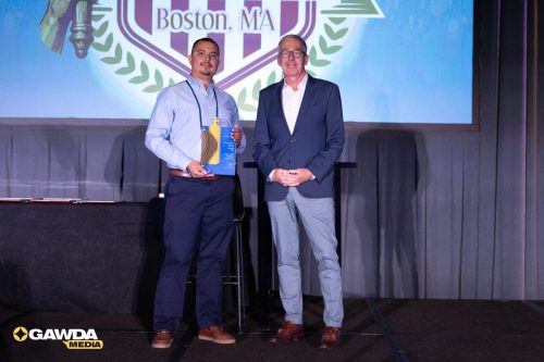 Two men on a stage, one holding an award plaque and bottle, with a Boston, MA logo backdrop behind them.