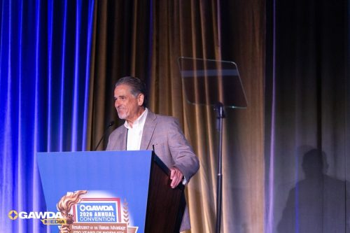 Man in a light gray blazer speaks at a blue podium on a stage with blue and gold curtains; podium displays the GAWDA 2026 Annual Convention logo and slogan headings.