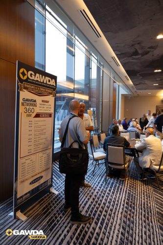 A conference lobby with a tall GAWDA banner and attendees seated around tables; a man with a backpack stands near the banner.