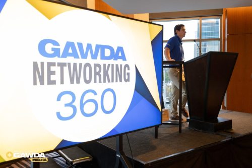 Speaker at a podium on stage during the GAWDA Networking 360 event, with a large branded display behind.