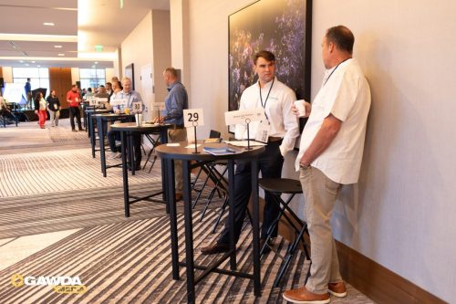 Conference hallway with round high-top tables and attendees standing and talking; table signs show 29–31; GAWDA Media logo bottom-left corner.
