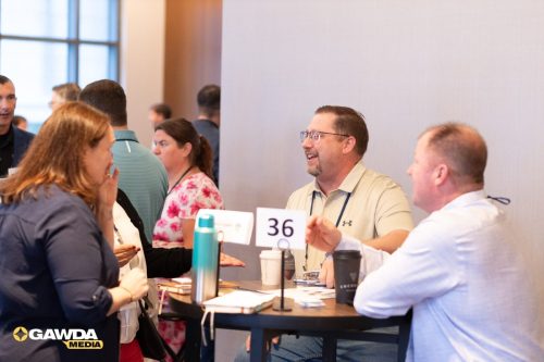 Professionals at a registration desk chatting, with a '36' sign on the table and coffee cups nearby.