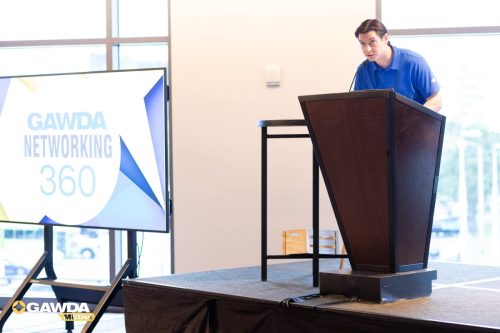 Man in a blue polo standing at a dark wooden podium giving a presentation, with a large screen showing 'GAWDA Networking 360' nearby.