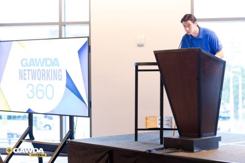 Man in a blue polo speaks from a dark wooden podium at a networking event, with a large screen reading 'GAWDA Networking 360' nearby.