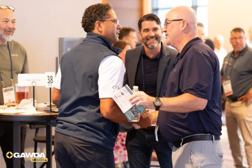 Two men shake hands at a conference, one holding a brochure and water bottle as others mingle in the background.