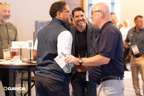 Two men shake hands and chat at a conference booth while a third man smiles in the background.