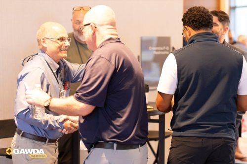 Two men shake hands and smile at a conference, one in a light blue shirt with a badge and a water bottle nearby.