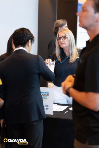 Woman with glasses and a name badge helping a man at a conference check-in desk; GAWDA logo visible in the corner.