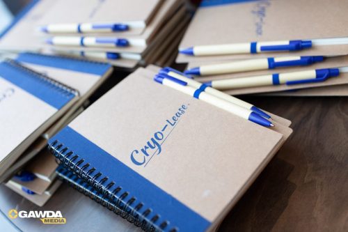 Stacks of brown and blue notebooks with blue pens scattered on a wooden desk; cover text reads 'Cryo-Lease' and GAWDA Media logo visible in the corner.