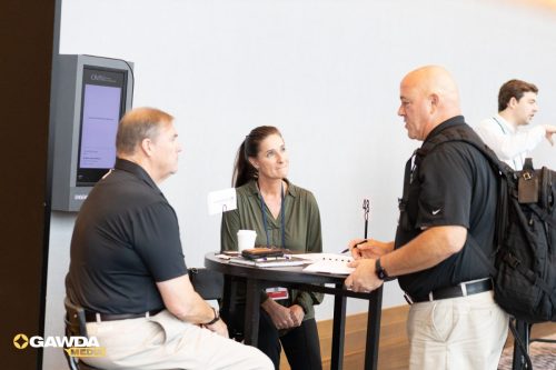 Three professionals in a conference setting gather around a tall round table, chatting; badges and papers on the table, GAWDA Media logo bottom left.