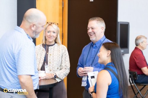 Group of professionals networking and chatting at a conference, smiling and holding name badges near a tall table. GAWDA Media logo visible in the corner.