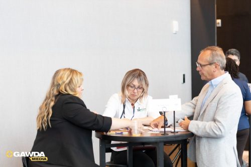 Three people sit around a high round table, a woman in a white shirt assists a man in a light blazer as another woman watches nearby lightheartedly.