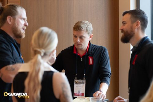 Man in a black shirt with a red lanyard badge labeled Cameron Sallee converses with three people at a table, GAWDPA Media logo visible in the corner.