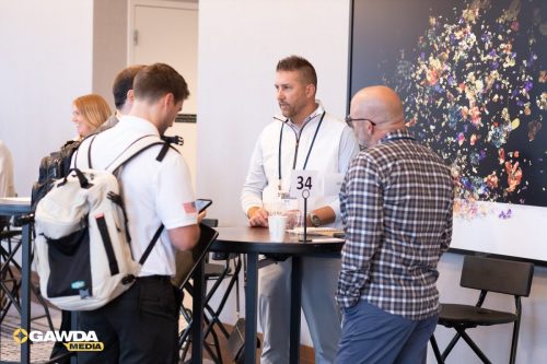 Group of professionals conversing around a high round table at a conference registration area, man with badge '34' speaking to attendees while a floral mural sits behind them.