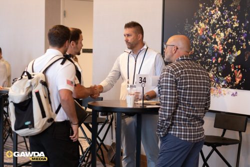Man in a light vest shakes hands with a man at a high round table during a conference, table number 34 visible, GAWDA Media logo in corner.