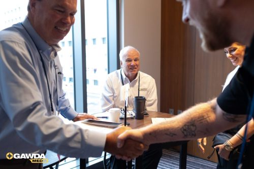 Three colleagues greet each other with a handshake across a table in a bright office, all smiling nearby Coffee cups and papers on the table suggest a meeting state of collaboration.