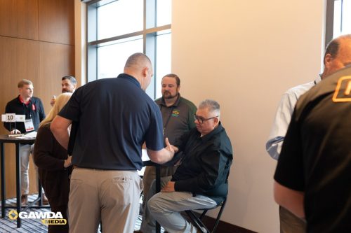 Professionals mingle at a conference registration area by windows; a man in a black shirt speaks with a seated attendee being assisted.