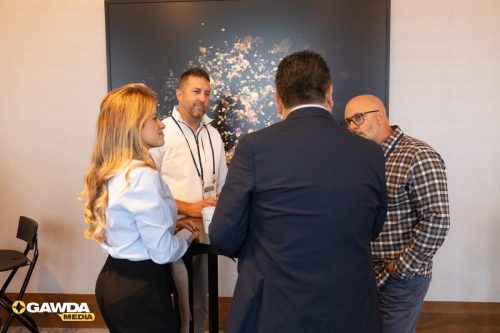Four professionals stand around a tall round table in a conference room, engaged in conversation with a floral artwork on the wall behind them. Contains a GAWDA Media logo in the bottom-left corner.