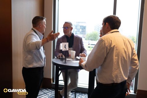Three businessmen stand around a high round table by a large window, mid-conversation; a '4' sign, coffee cups, and name badges are visible.