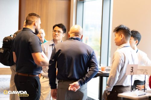 Group of professionals in a business casual setting chat around a standing table by a window at a conference, with a '3' sign on the table and GAWDA Media logo visible in the corner.