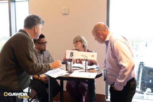 Four professionals sit and stand around a round table in a bright office, reviewing papers; a sign with '8' and 'AWISCO' sits on the table with coffee cups nearby.