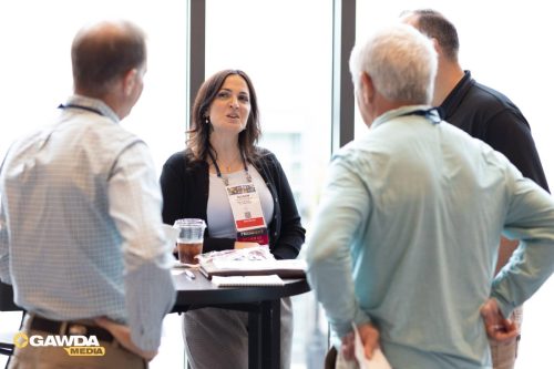 Woman with a conference badge talking to three men around a high-top table by a bright window.