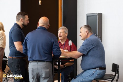 Group of men in polo and polo shirts chat around a high table at a conference area, GAWDA MEDIA logo visible in the corner.