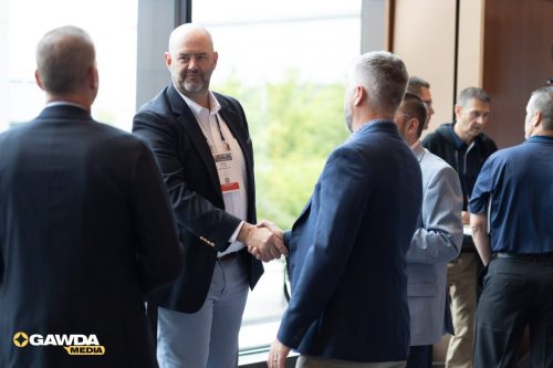 Two men in suits shake hands at a conference, while other attendees chat near a window; conference badges visible on lanyards.