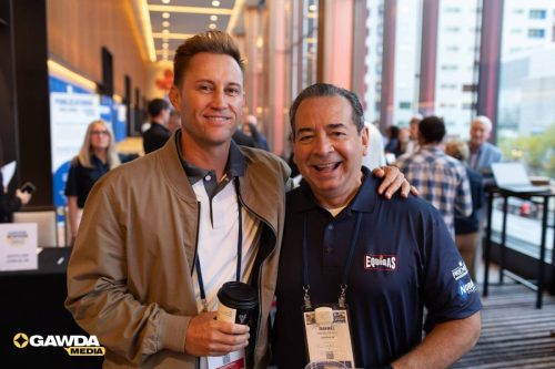 Two men pose for a photo at a conference lobby; one in a beige jacket holding a coffee cup, the other in a navy polo with a name badge.