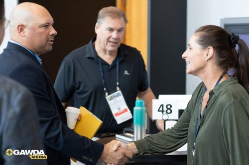 Two professionals shake hands at a conference check-in desk, with a staff member in the background.