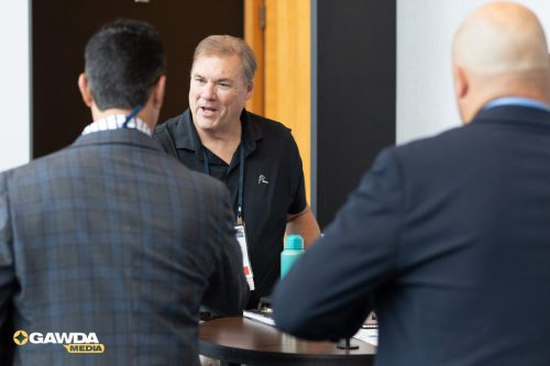 A man in a black polo talks with two attendees at a round table during a conference; GAWDA Media logo in the bottom-left corner.