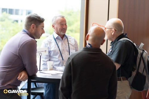Four men stand around a tall table at a bright conference space, chatting and smiling, with a GAWDA sign nearby. (Note: GAWDA logo visible in the corner.)