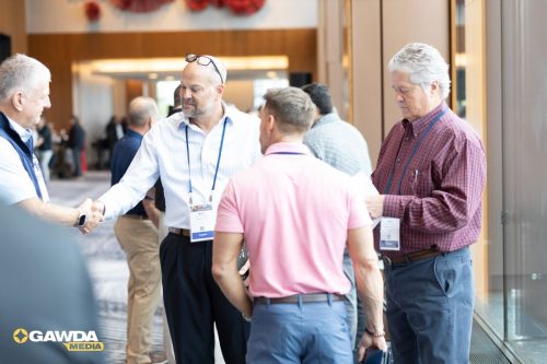 Men in a conference lobby shaking hands and networking, with name badges visible under bright indoor lighting.