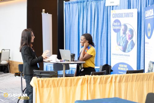 Two professionals converse at a booth: a standing woman with papers and a smiling seated woman with a laptop, blue backdrop behind them.