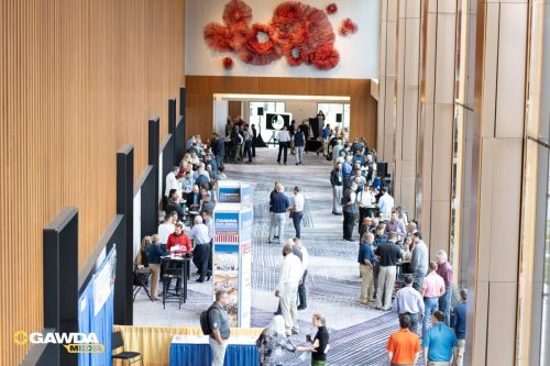 Crowded conference lobby with attendees networking near exhibitor booths and a stage in the back.