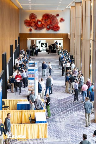 Crowded conference hall with attendees around exhibitor booths along a patterned carpet, a bright orange flower sculpture on the back wall visible in the background, and a GAWDA Media logo in the corner.