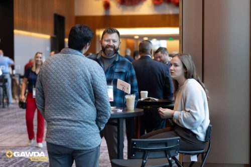 Smiling man in a blue plaid shirt talks with attendees at a standing networking table during a conference; coffee cups sit on the table.