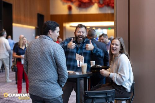 Three people at a standing round-table networking area: a bearded man in a plaid shirt gives two thumbs up while a smiling woman sits nearby with coffee cups on the table behind them.
