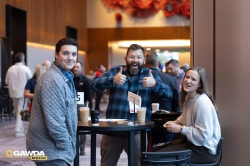 Three people at a high-top conference table pose for a photo, drinks on the table and a GAWDA Media logo visible in the corner, smiling at the camera.