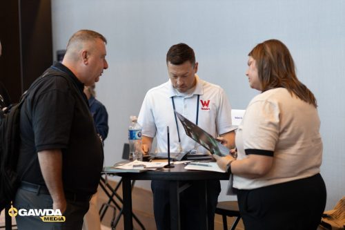 Three people stand around a high round table at a trade show, examining brochures; the man in the center wears a white polo with a red Welder’s logo. A water bottle and scattered papers sit on the table.