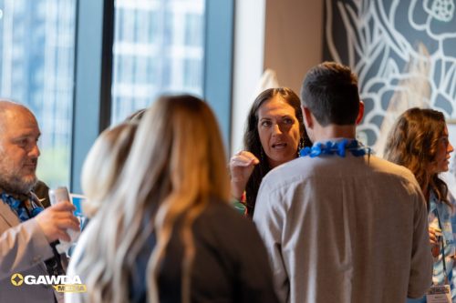 Woman speaks to a small group at a networking event, gesturing with her hand while attendees listen in a bright room. A man in the foreground wears a blue lei around his neck.