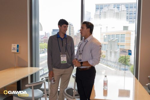 Two professionals with ID badges chatting in a bright office by a large window overlooking a cityscape, with a bottle of water on the table nearby.