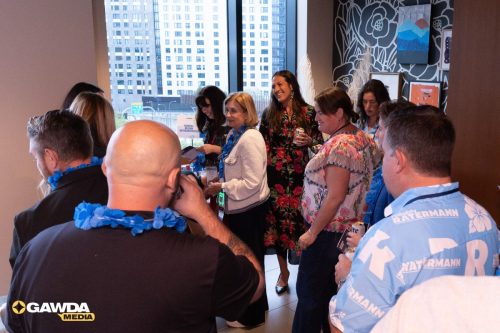 Group of adults at a social/corporate event in a bright office, wearing blue leis and name badges, with city buildings visible through the windows.