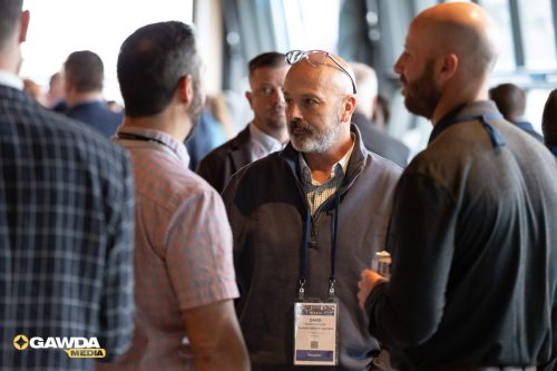 Groups of men networking at a conference; man in center wears glasses on his head and a name badge reading DAVID talking with others.